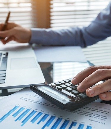 Männliche Person im Business Look an einem Schreibtisch mit Laptop, Taschenrechner und DOkumente mit Diagrammen