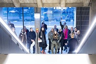 Eine Gruppe von Menschen hört einem Vortragenden in einem modernen Gebäude zu, während sie auf einer Treppe oder Plattform stehen; im Hintergrund sind große Fenster mit Blick auf die Straße.