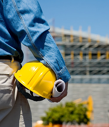 Person mit gelben Bauhelm blickt auf eine großes Gebäude, deren Fassade saniert wird