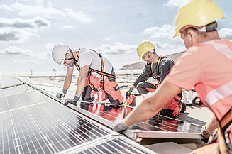 Zu sehen sind drei Baustellenmitarbeiter die gerade eine Photovoltaik-Anlage auf einem Bauen im Bestand-Projekt montieren