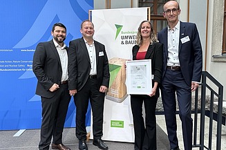 Gruppenfoto: 3männliche Personen, eine weibliche Person, stehend vor einem Roll Up mit der Aufschrift UMWELT & BAUEN Bundespreis. Sie halten eine Urkunde in der Hand