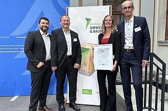 Gruppenfoto: 3männliche Personen, eine weibliche Person, stehend vor einem Roll Up mit der Aufschrift UMWELT & BAUEN Bundespreis. Sie halten eine Urkunde in der Hand