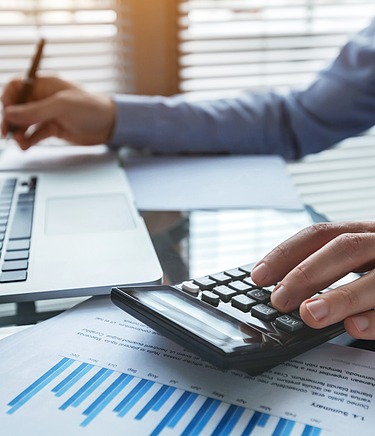 Männliche Person im Business Look an einem Schreibtisch mit Laptop, Taschenrechner und DOkumente mit Diagrammen