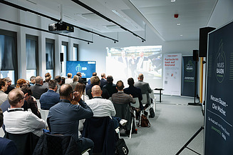 Fachforum mit Zuhörern die auf eine Leinwand schauen mit einem Banner zum Thema Bauen im Bestand