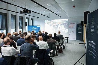 Fachforum mit Zuhörern die auf eine Leinwand schauen mit einem Banner zum Thema Bauen im Bestand