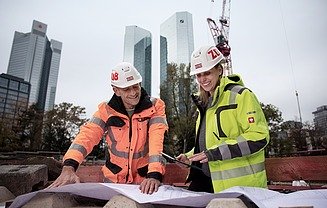 Projektmitarbeiter lesen Baulpläne auf einer Baustelle im Hintergrund sind Wolkenkratzer und ein Krahn