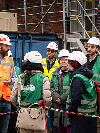 Detailaufnahme Baustellenbesichtigung Personengruppe und ein Gruppenführer der nach  oben zeigt, alle tragen Bauhelme