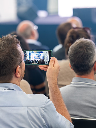 Teilnehmer von hinten nimmt mit seinem Handy die Podiumsdiskussion auf