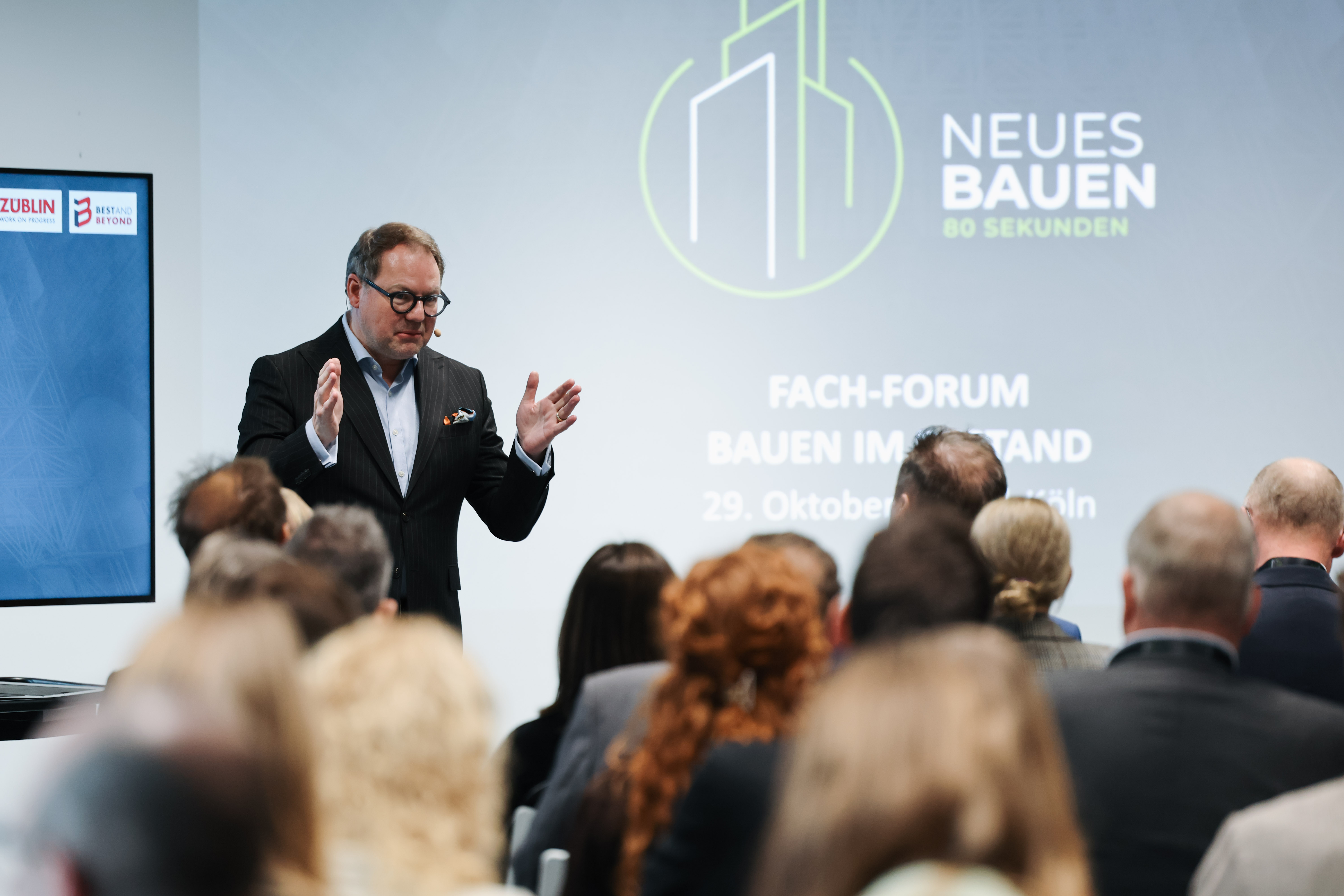 Ein Vortragsredner vor einer Gruppe, im Hintergrund an der Leinwand Neues Bauer - 80 Sekunden