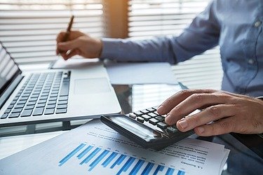 Männliche Person im Business Look an einem Schreibtisch mit Laptop, Taschenrechner und DOkumente mit Diagrammen