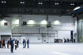 Eine Gruppe von Menschen befindet sich in einer großen, leeren Industriehalle mit hohen Decken und weißen Backsteinwänden; einige Personen stehen zusammen, andere gehen in einer Reihe durch ein großes Tor.