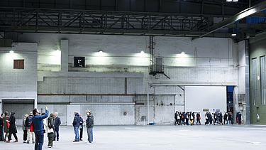 Eine Gruppe von Menschen befindet sich in einer großen, leeren Industriehalle mit hohen Decken und weißen Backsteinwänden; einige Personen stehen zusammen, andere gehen in einer Reihe durch ein großes Tor.