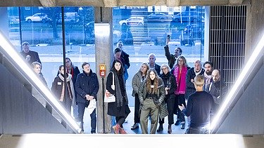 Eine Gruppe von Menschen hört einem Vortragenden in einem modernen Gebäude zu, während sie auf einer Treppe oder Plattform stehen; im Hintergrund sind große Fenster mit Blick auf die Straße.