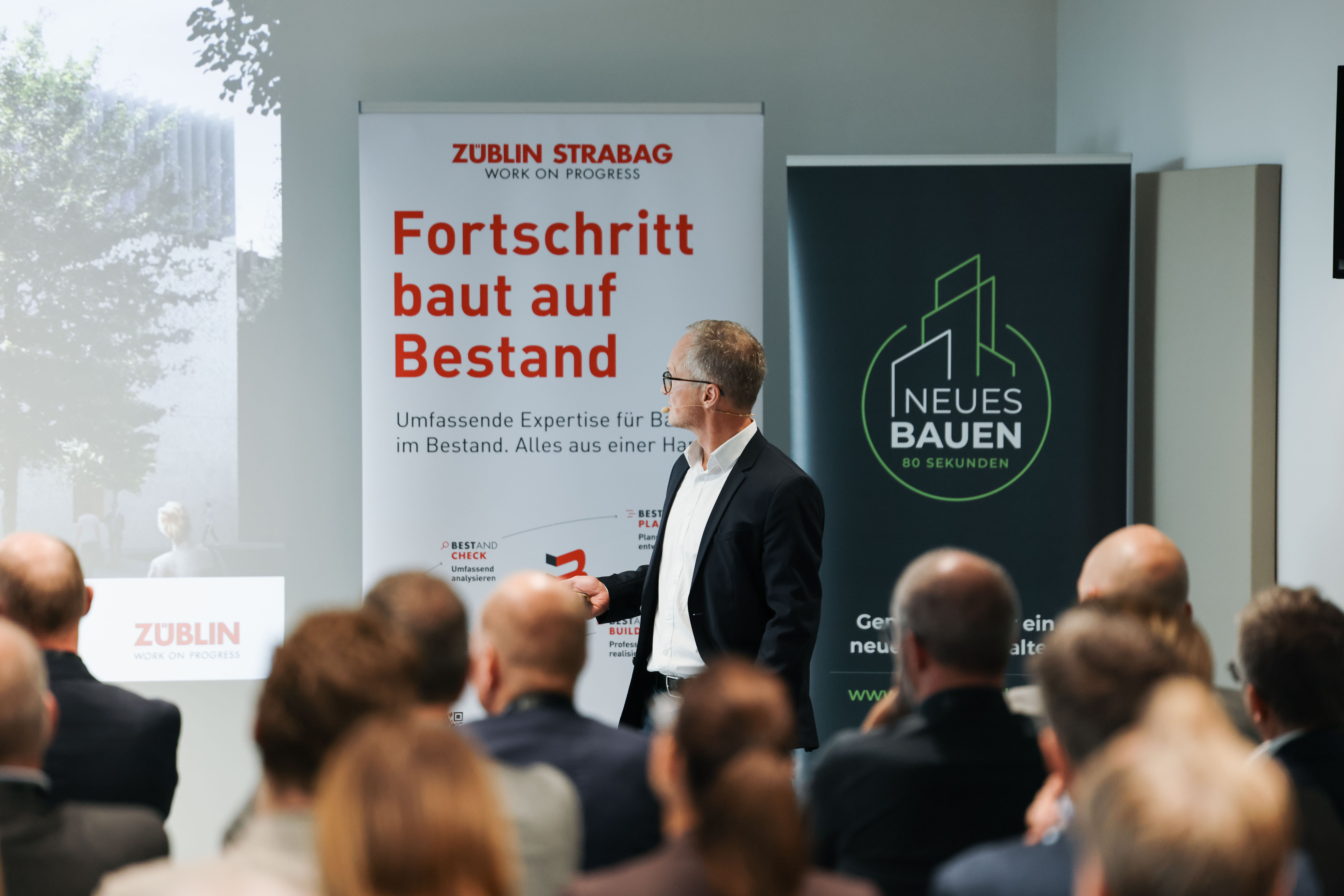 Vortragsredner vor einer Gruppe. Im Hintergrund auf der Leinwand eine ZÜBLIN Präsentation und ein Banner mit der Botschaft Fortschrift baut auf Bestand