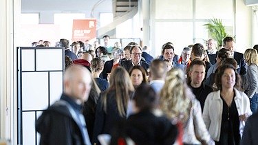 Viele Menschen bewegen sich durch einen hellen Innenraum mit offenen Strukturen und einem Spiral-Treppenaufgang im Hintergrund; es wirkt wie ein lebhaftes Networking-Event.