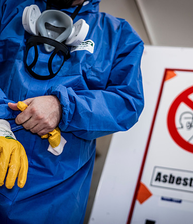 Person in Schutzkleidung dahinter ein Gefahrenschild mit der Aufschrift 