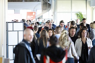 Viele Menschen bewegen sich durch einen hellen Innenraum mit offenen Strukturen und einem Spiral-Treppenaufgang im Hintergrund; es wirkt wie ein lebhaftes Networking-Event.